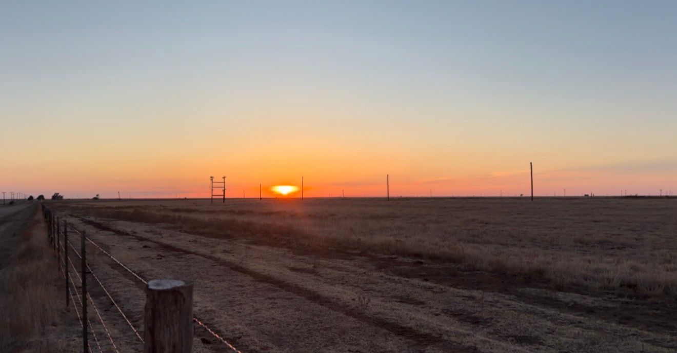 Golden sunset over flat farmland with a dirt road and fence line stretching toward the horizon under a vivid orange sky.