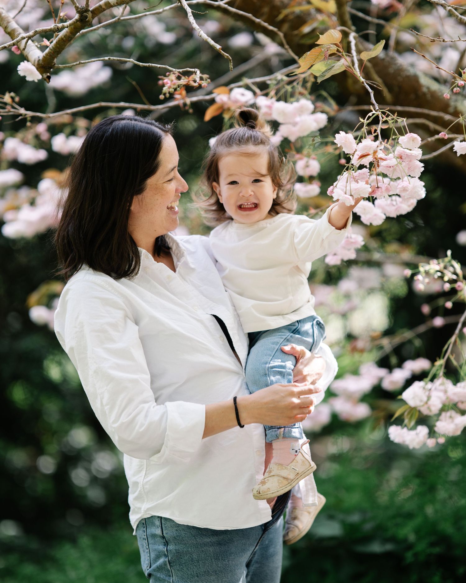 A smiling woman in white holds a laughing toddler reaching for pink cherry blossoms in a sunlit garden.