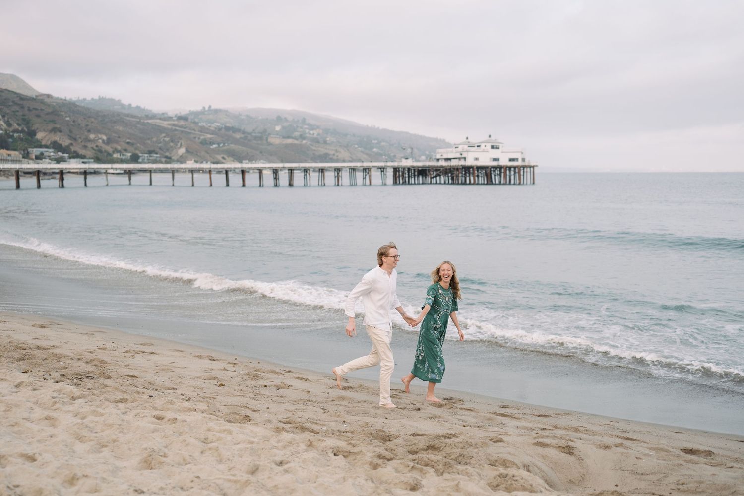 Couple holding hands running on Malibu beach with pier and ocean waves in background on overcast day.