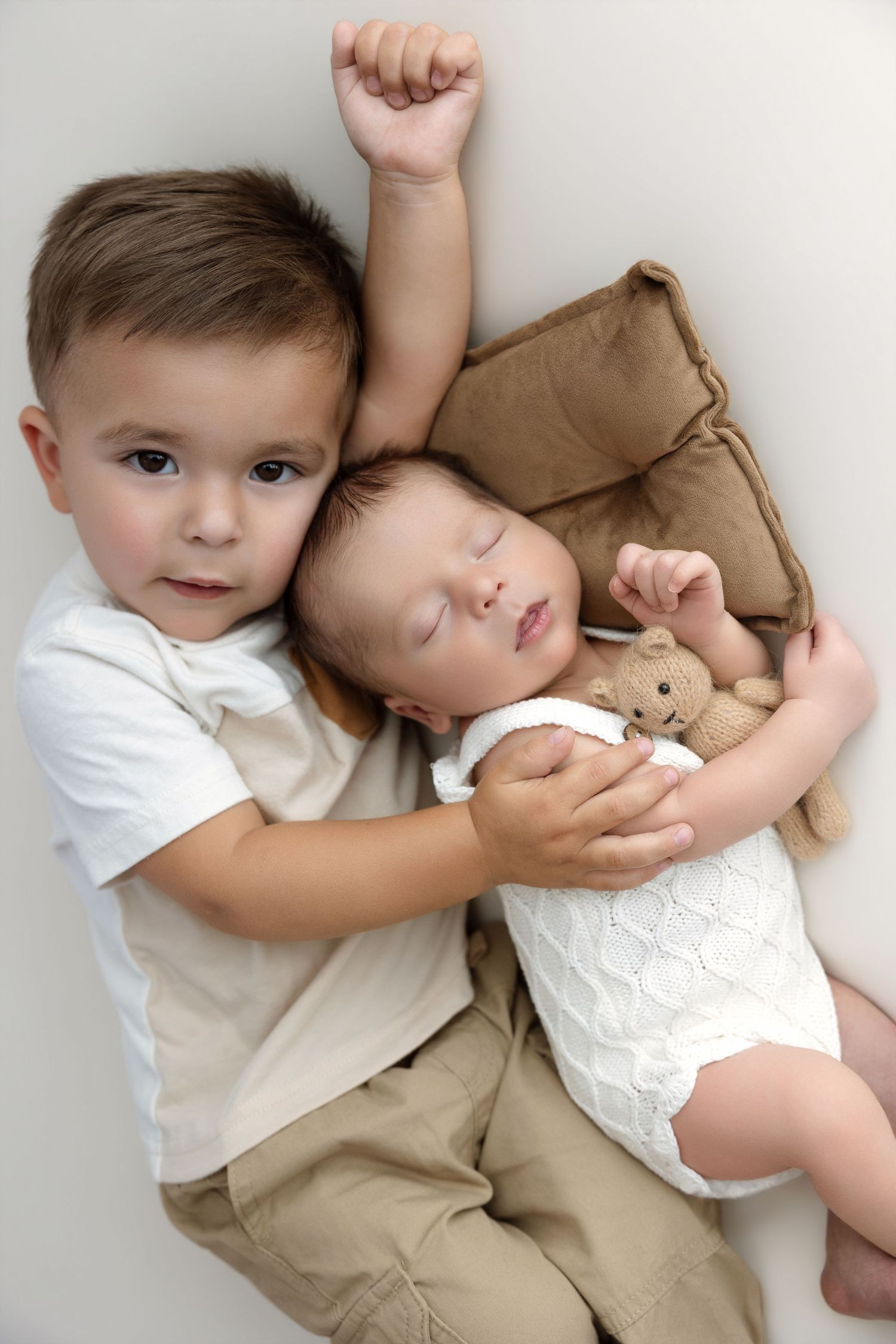 Young boy in white shirt gently holds sleeping newborn sibling with teddy bear on cozy brown couch.