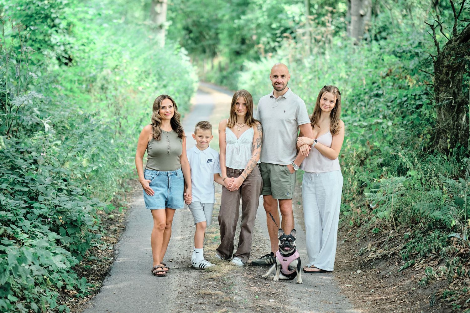 Family of five with a dog posing on a tree-lined path in a lush green forest setting.
