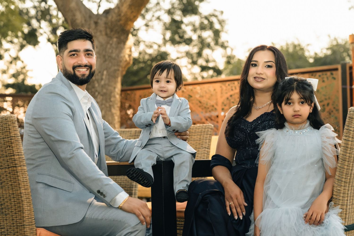 A smiling family of four poses outdoors, parents in formal attire with two young children dressed elegantly for a special occasion.