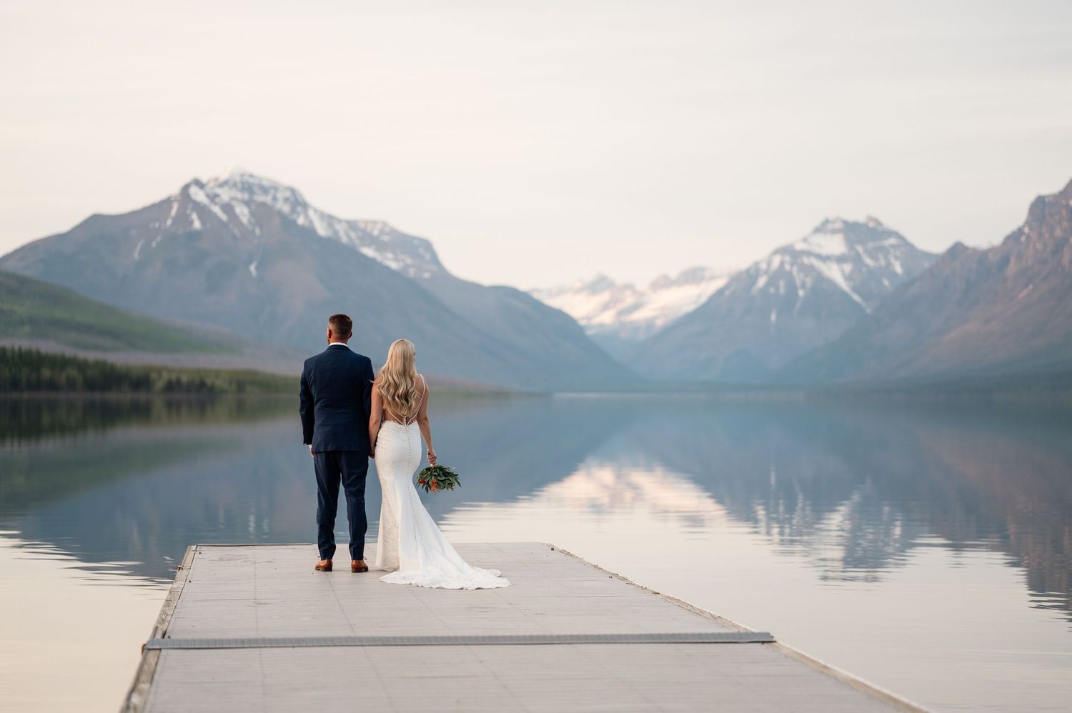 Bride and groom standing on a dock overlooking a serene mountain lake with snow-capped peaks at sunset.
