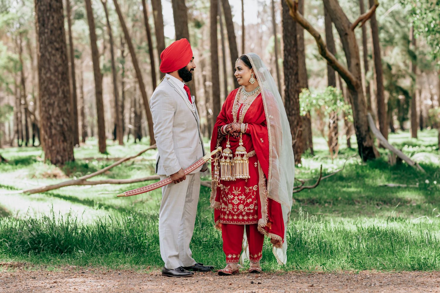 Sikh couple in traditional wedding attire standing in a forest, groom in white sherwani and red turban, bride in red salwar.