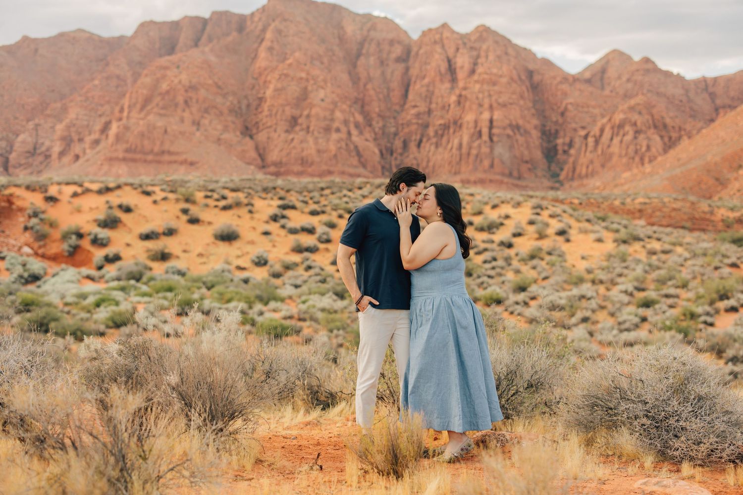 Couple sharing a romantic kiss in front of stunning red rock mountains in a desert landscape.