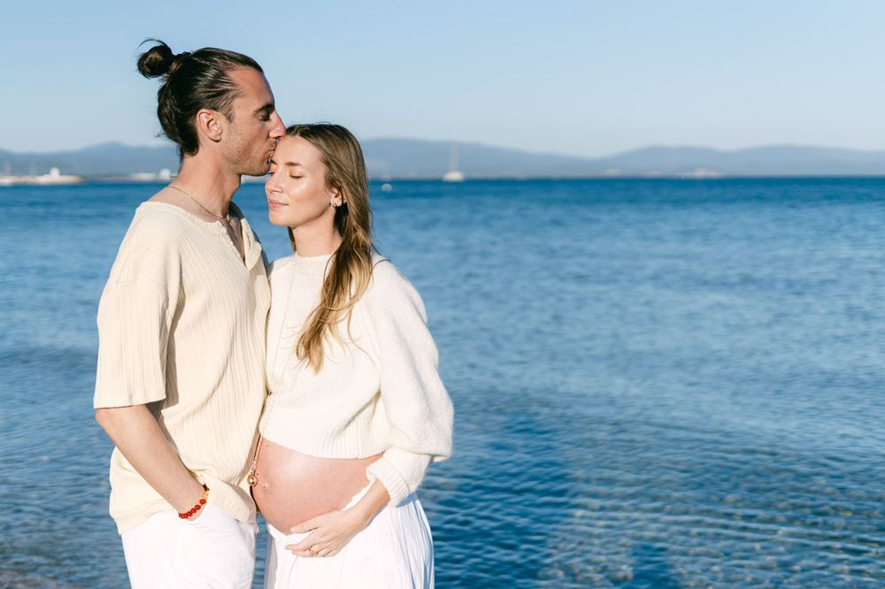 A couple in light colored clothing embraces by the blue ocean on a sunny day.