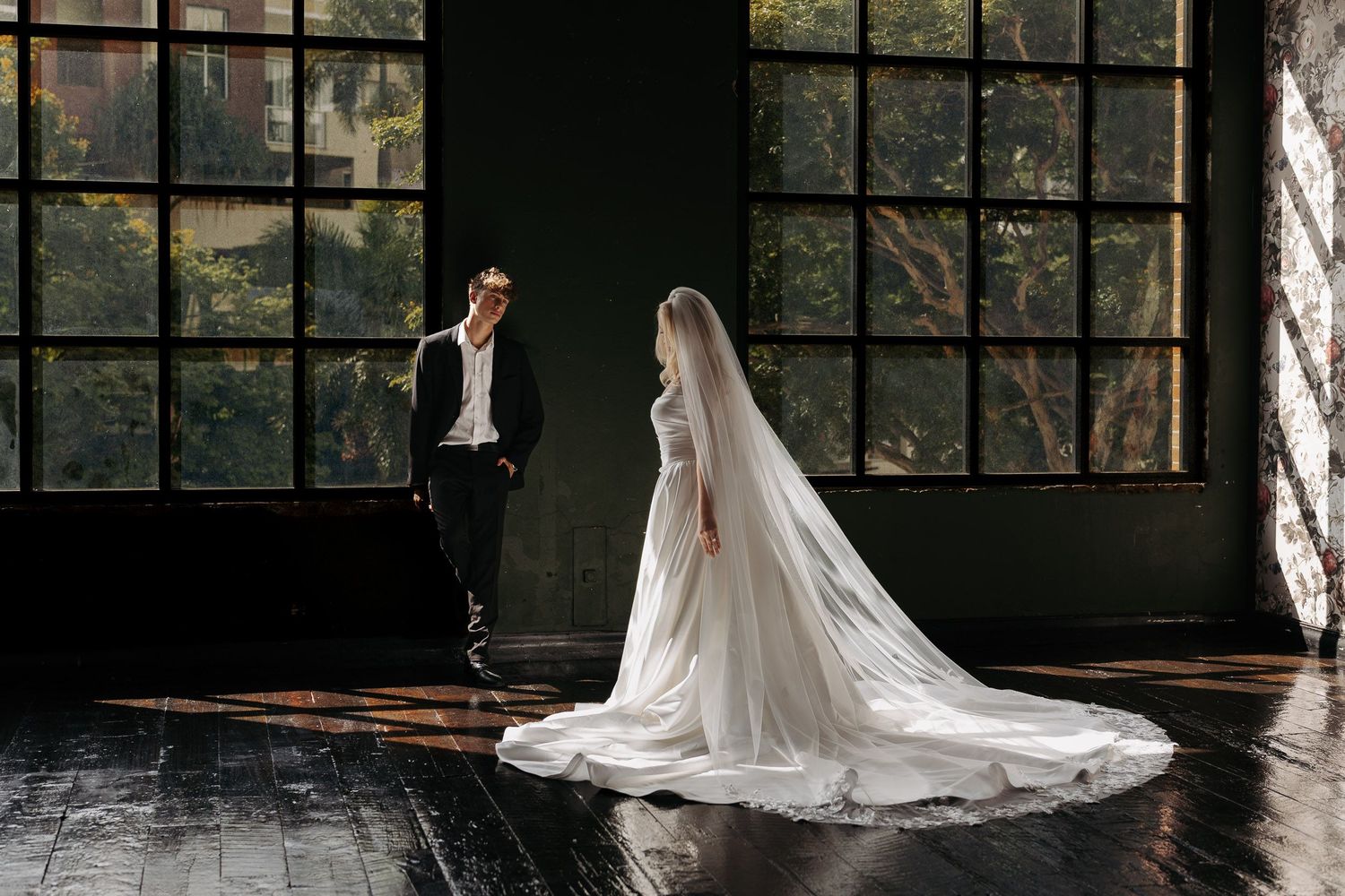 A bride in a flowing white gown with long veil stands near large windows while a groom leans against the wall.