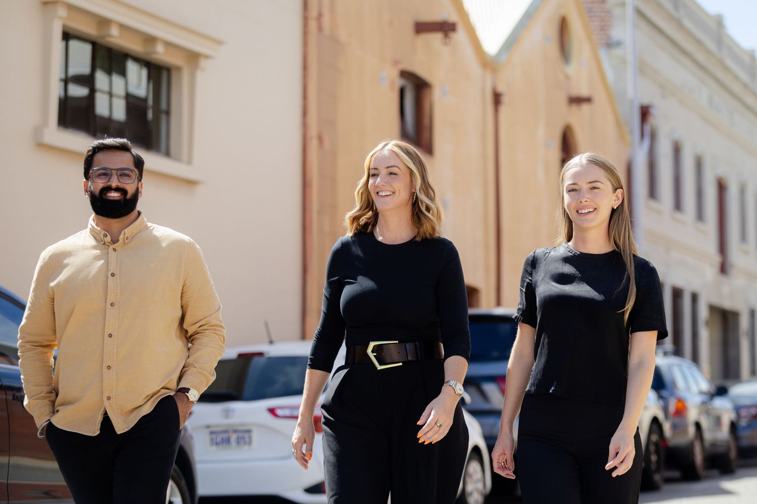 Three people walking together on a sunny street with parked cars and historic buildings in the background.