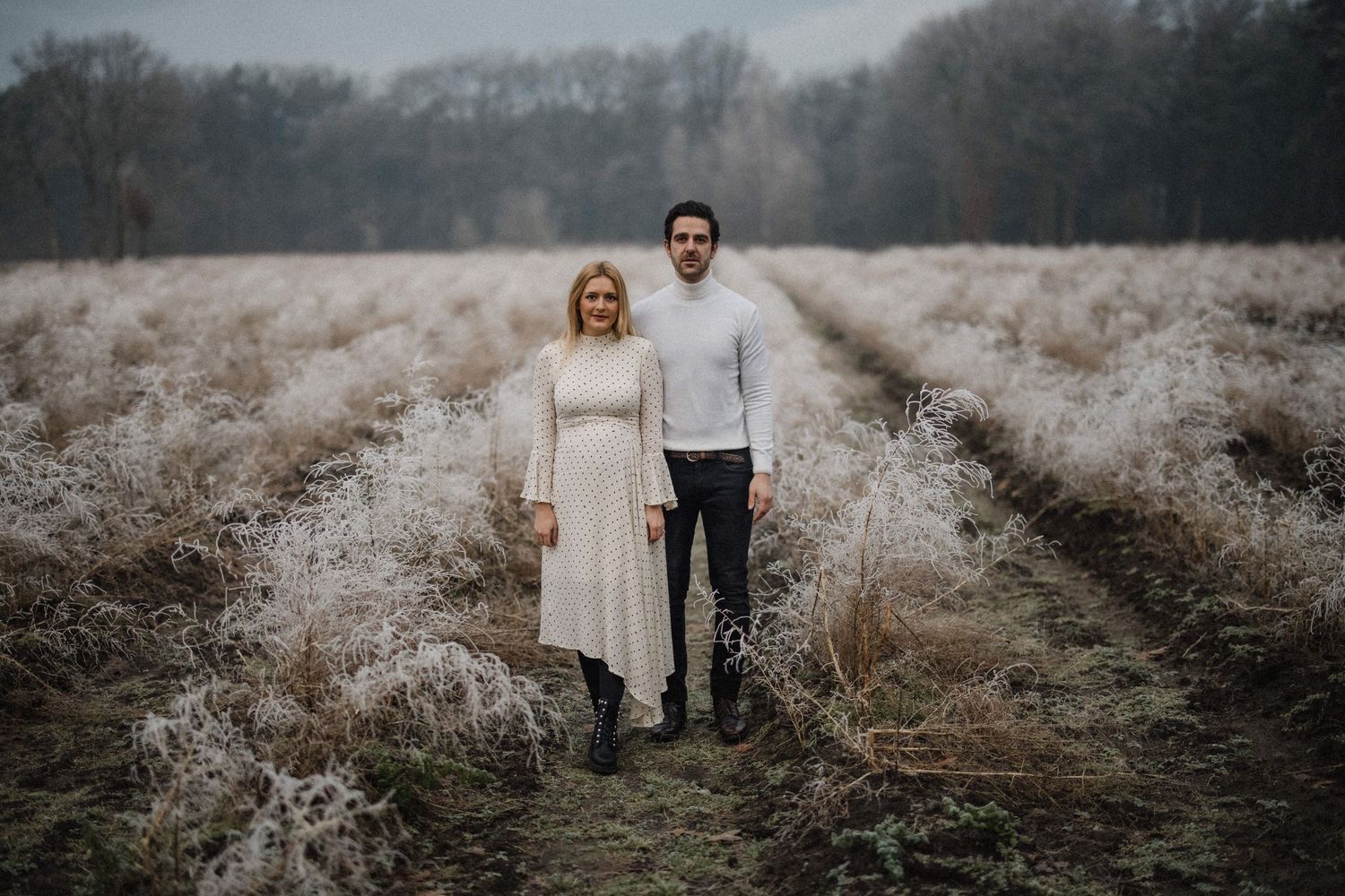 Een stel staat samen in een met vorst bedekt veld op een mistige winterochtend met kale bomen op de achtergrond.
