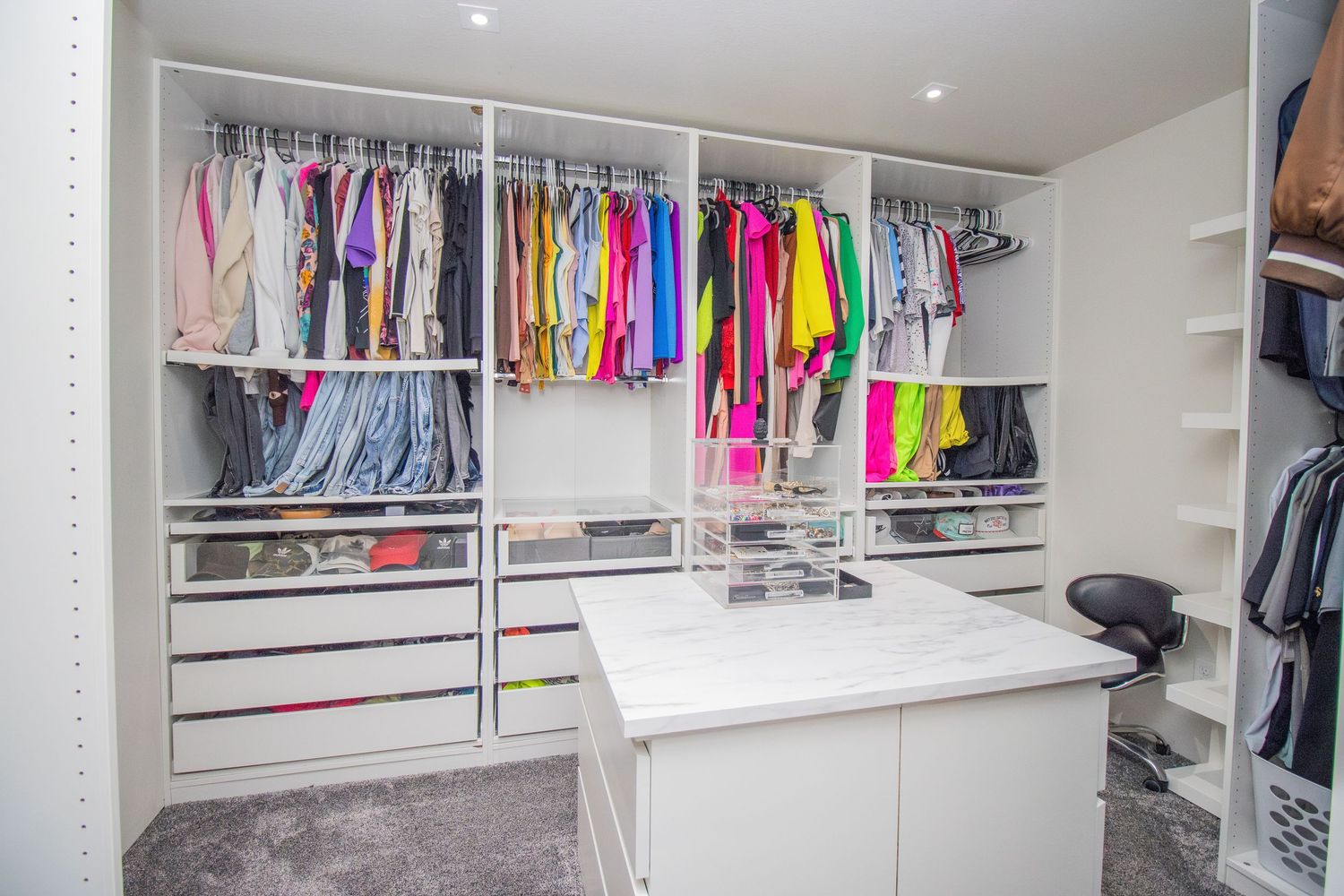 Spacious walk-in closet with white shelving, colorful hanging clothes, center island, and gray carpet flooring.