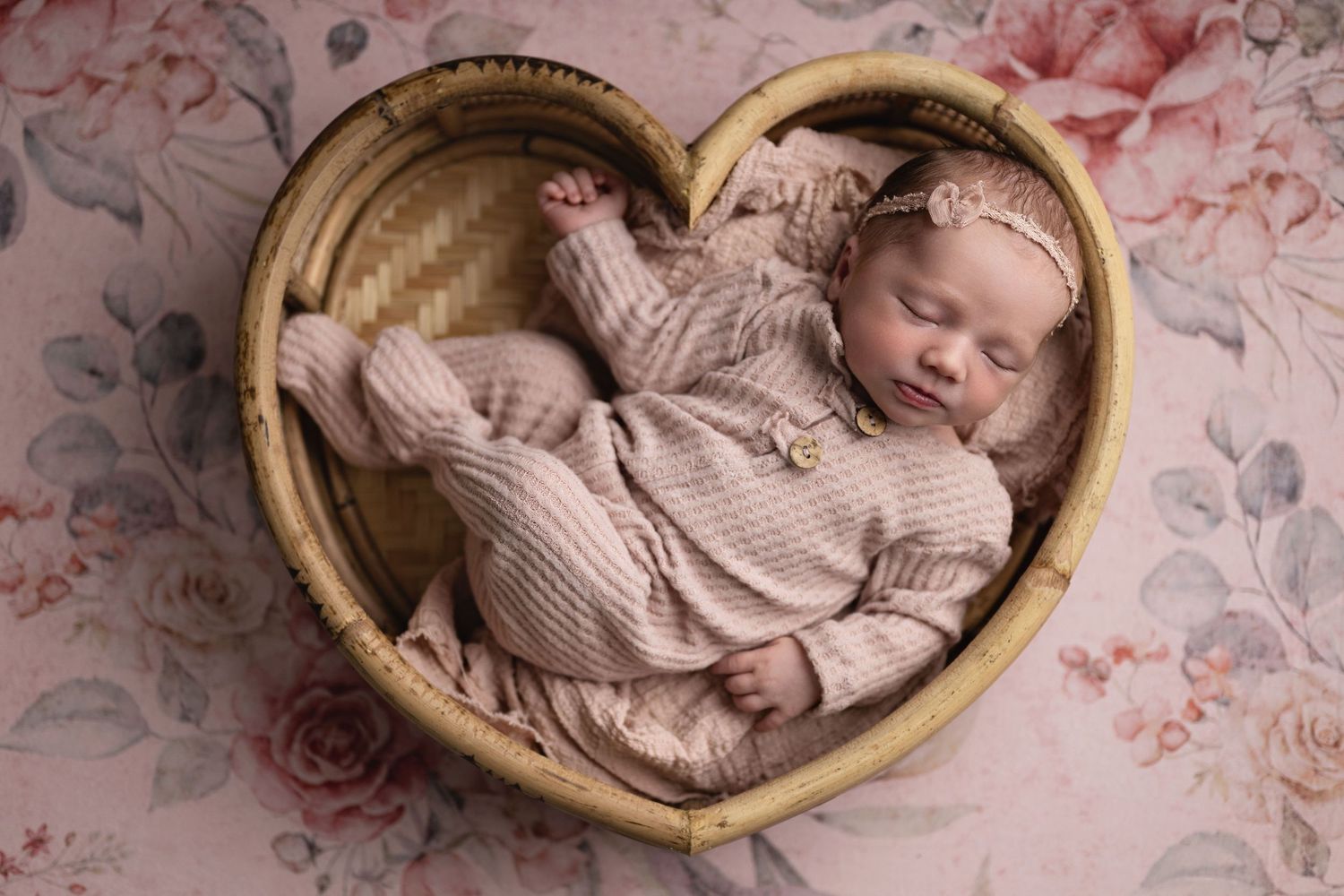 Sleeping newborn baby in pink outfit and headband resting in heart-shaped wicker basket on floral background.