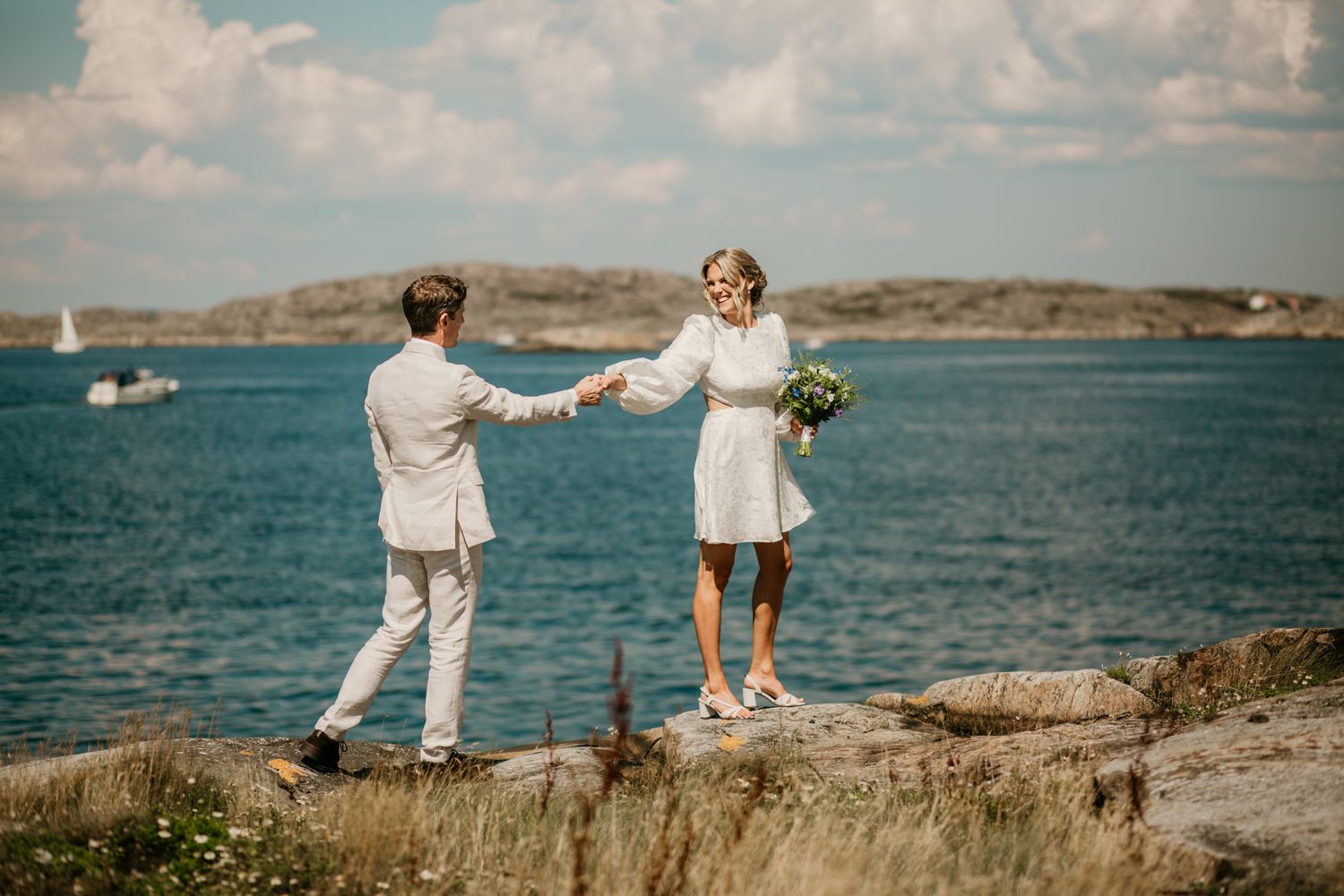 A couple in white outfits hold hands on a rocky coastal cliff with sailboats and blue water in the background.