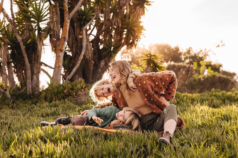 A mother and her two children laughing and playing together on green grass in a sunlit park with tropical trees.