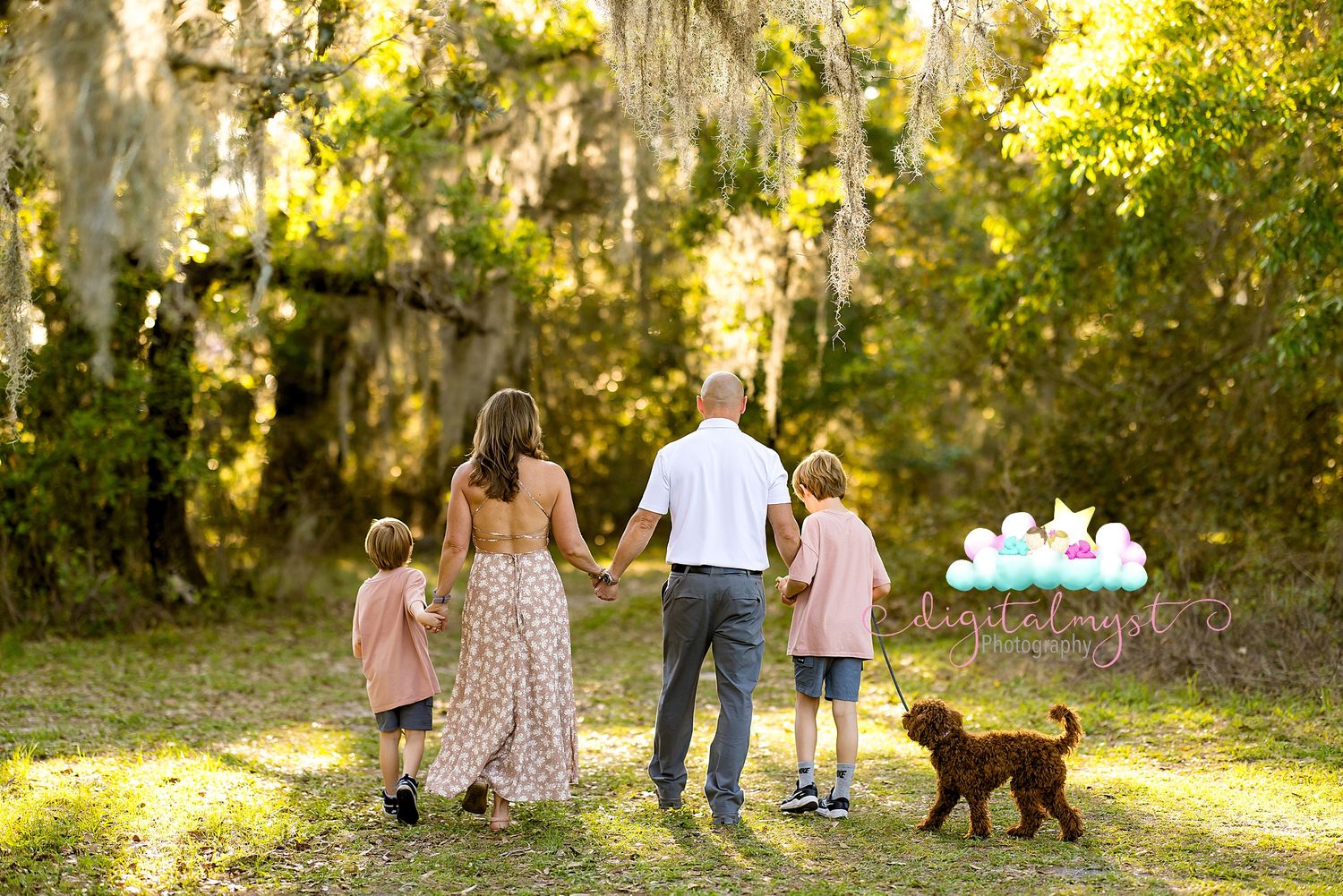 Family of four holding hands walking through a lush green park with their brown dog and balloons.