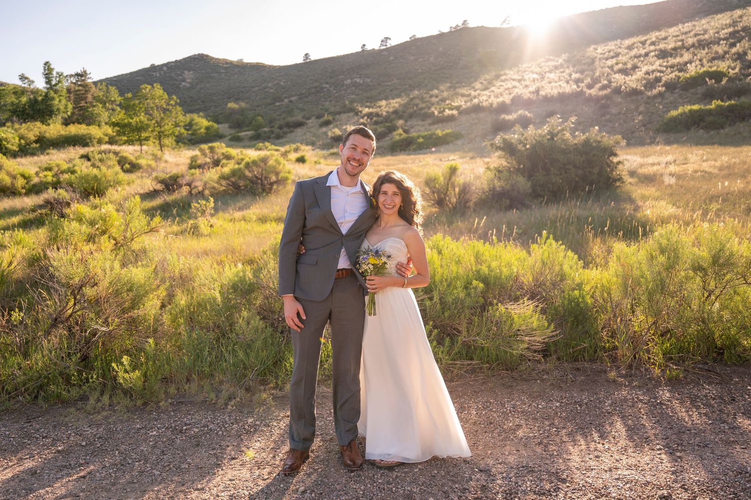 Bride and groom posing outdoors at sunset with green hills and wildflowers in a rustic mountain wedding setting.