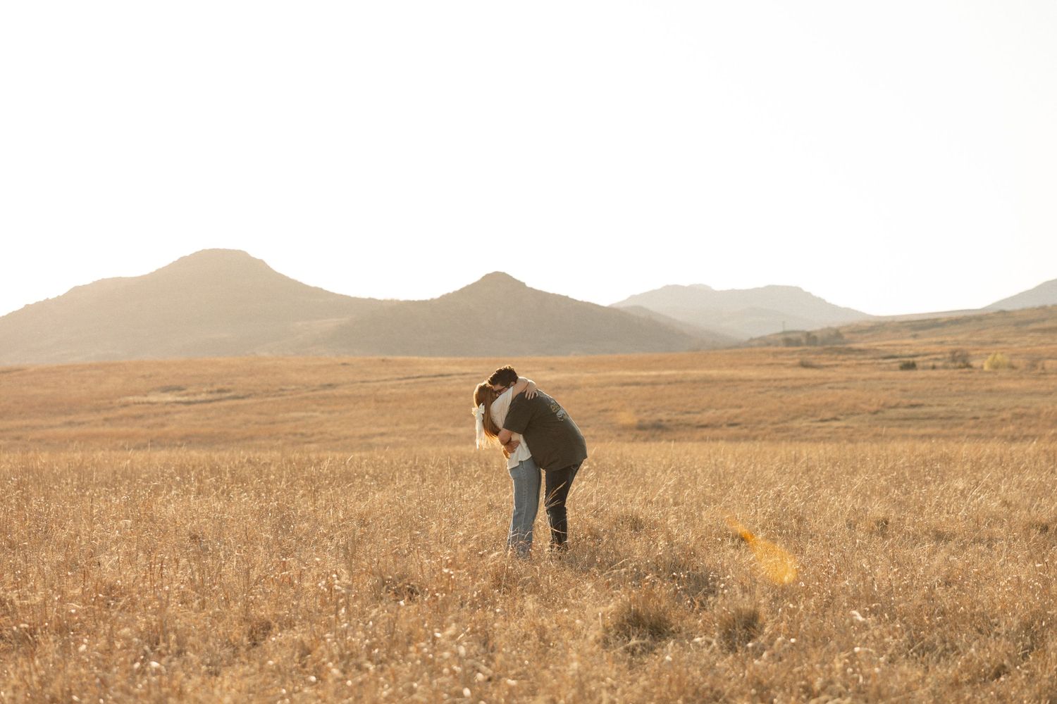 A person bends down in a golden field with mountains in the background at sunset.