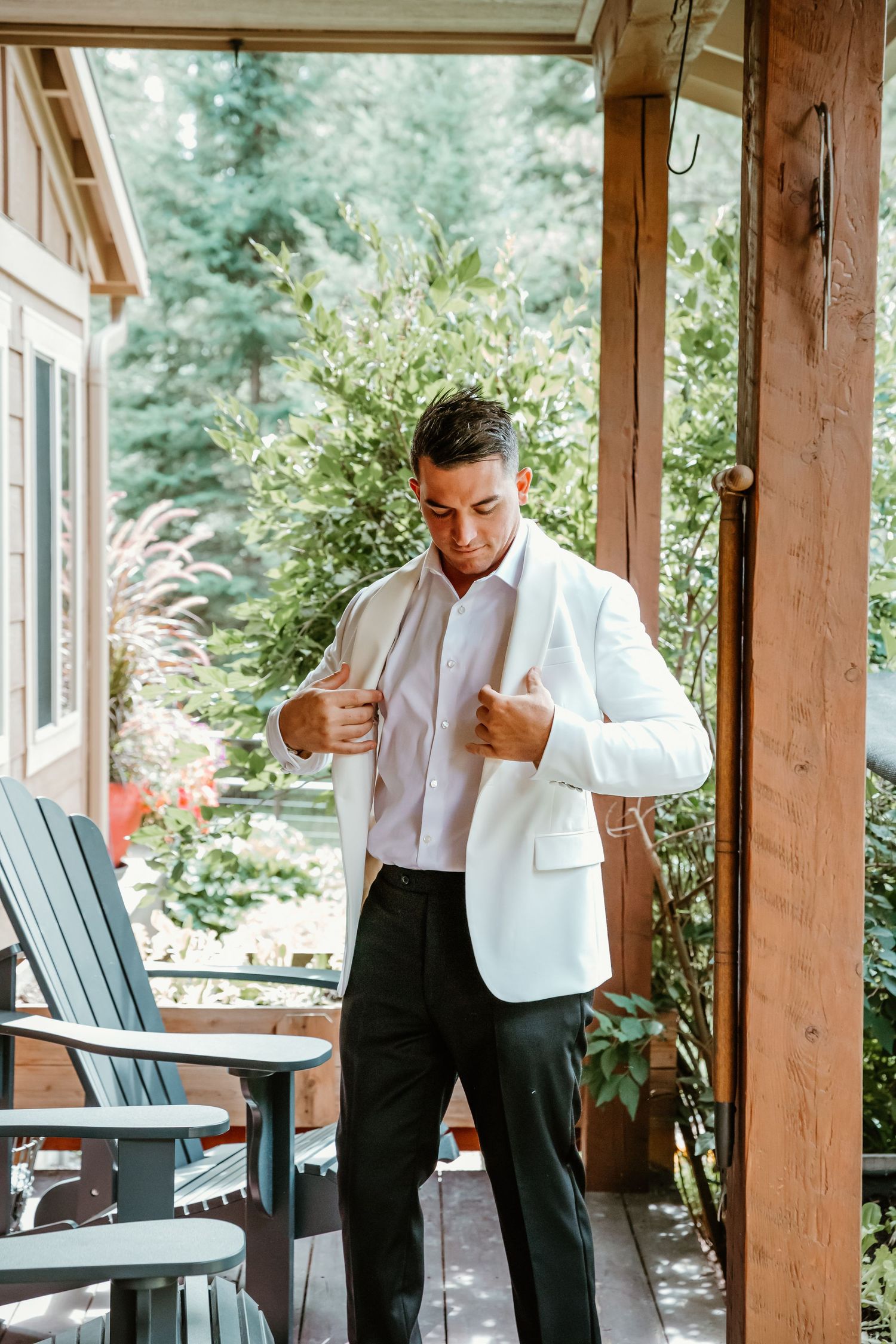 Groom adjusts white jacket while standing on wooden porch surrounded by greenery.