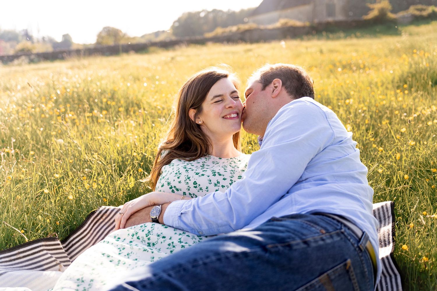 Man and woman lying on a blanket in a field of buttercups, sunset shining behind them. The man is embracing and kissing her on the cheek