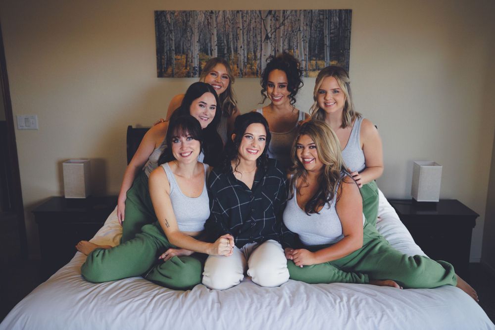 Seven women pose together smiling on a bed during a bridal party getting ready session.