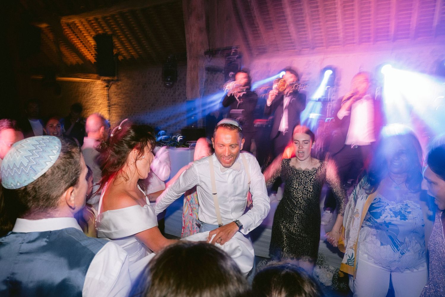 Wedding guests dance and celebrate on a lively dance floor illuminated by colorful party lights in a rustic venue.