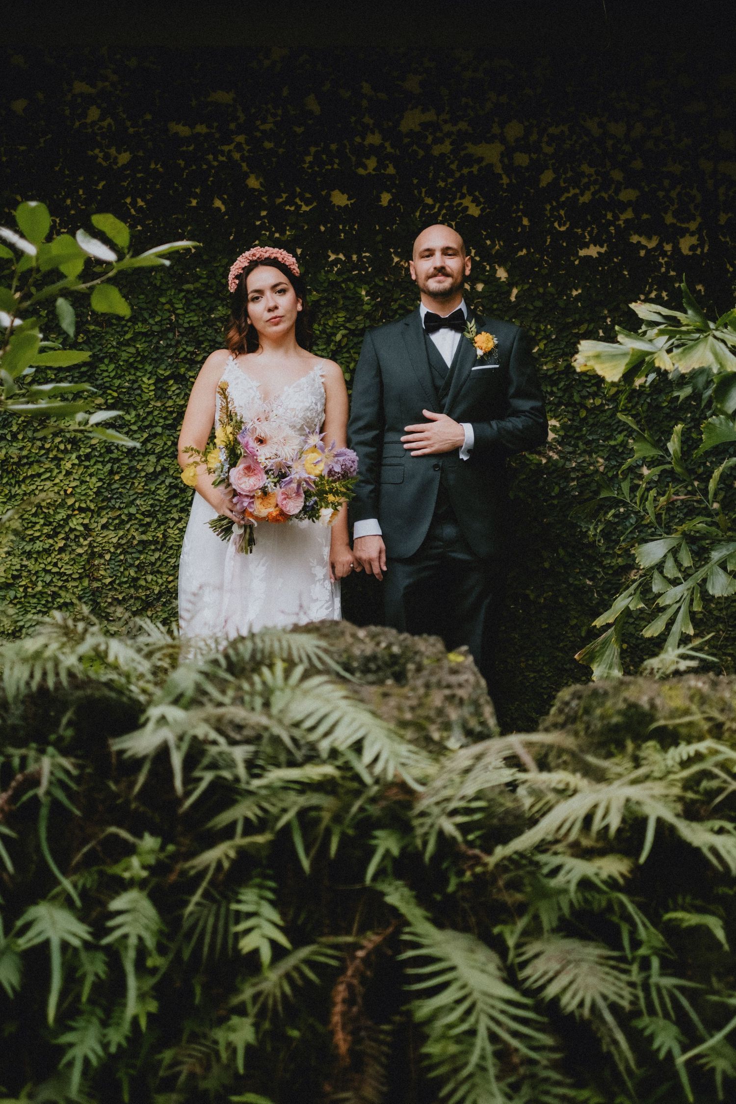 Couple stands together in lush garden setting wearing formal wedding attire surrounded by green ferns and foliage.