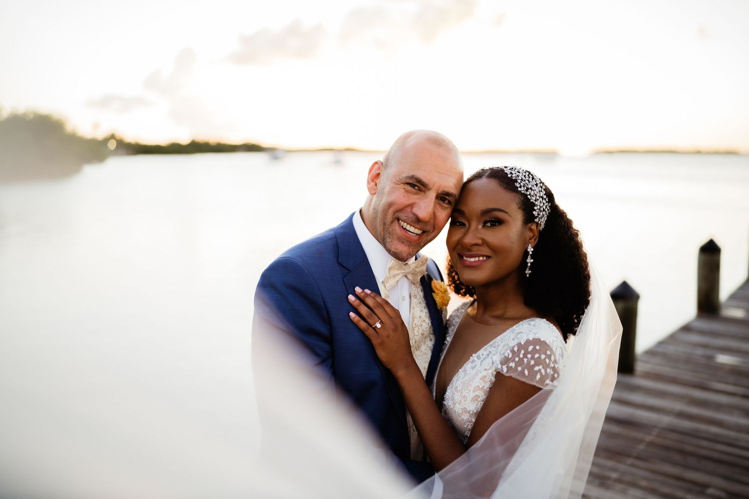 A romantic wedding couple shares intimate moments on a dock overlooking a serene waterfront at sunset.