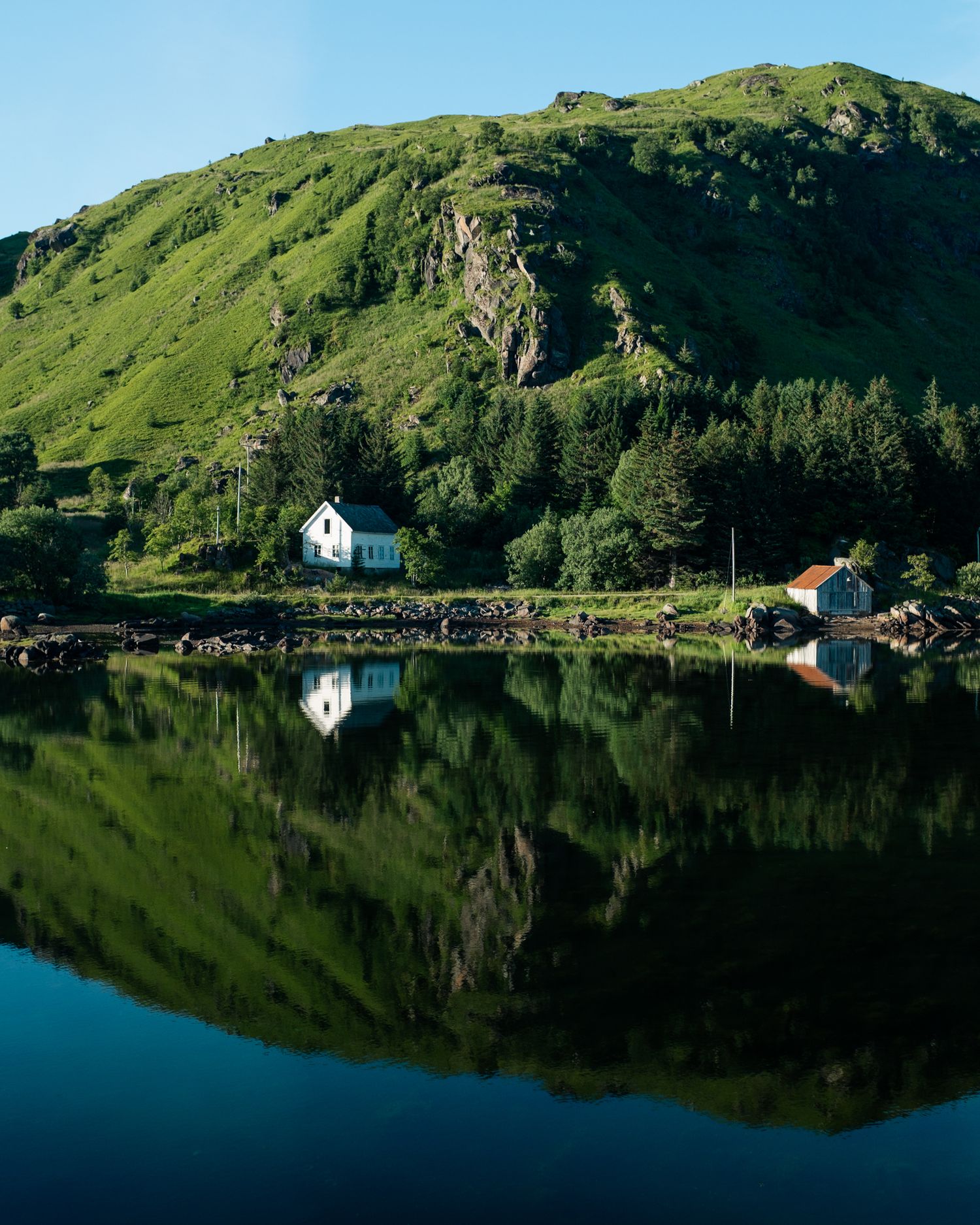 A small white church and buildings reflect perfectly in the still waters of a lake surrounded by green hills in Scotland.