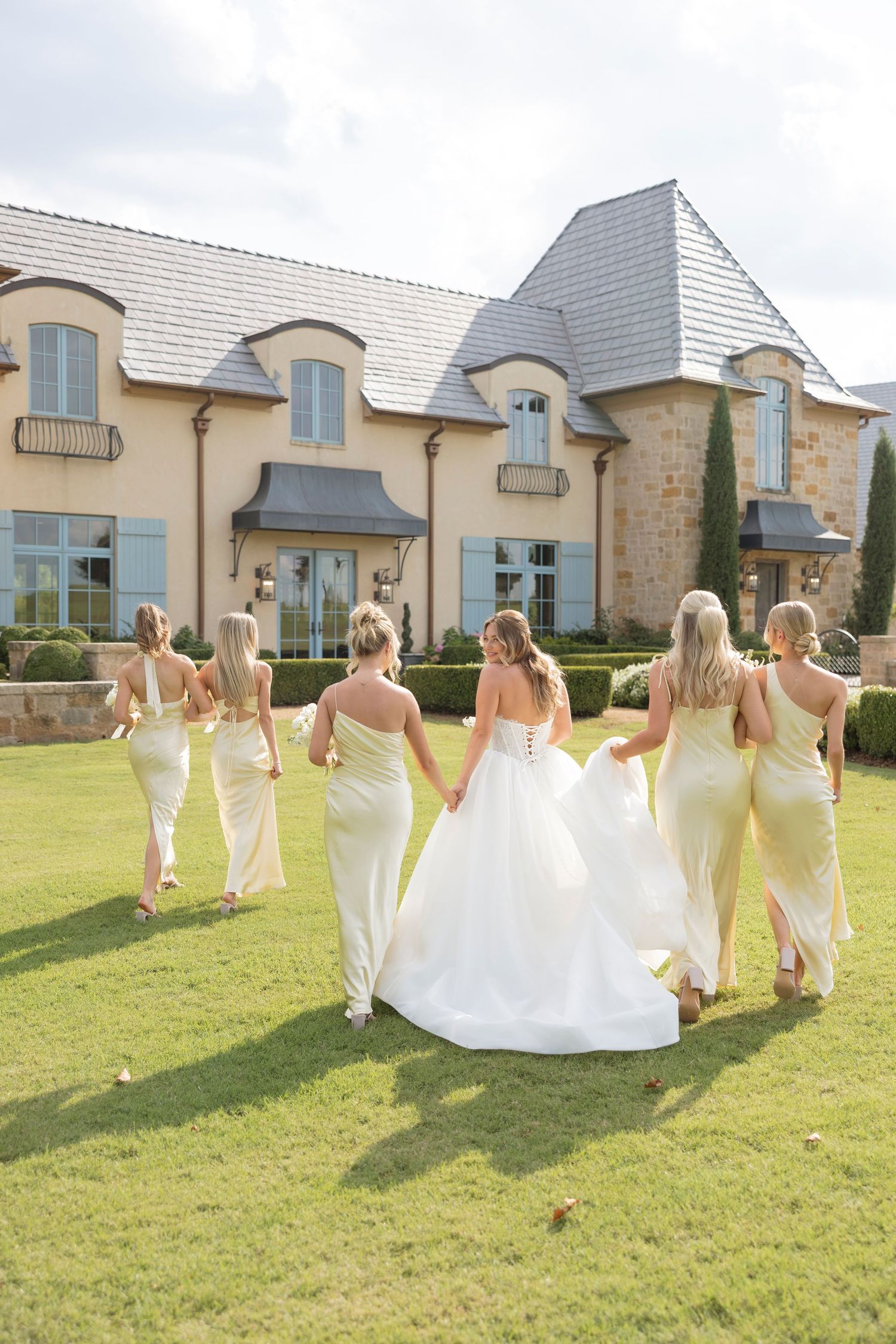 A group in yellow bridesmaid dresses and a bride in white walk together on a lawn in front of a French country-style mansion.