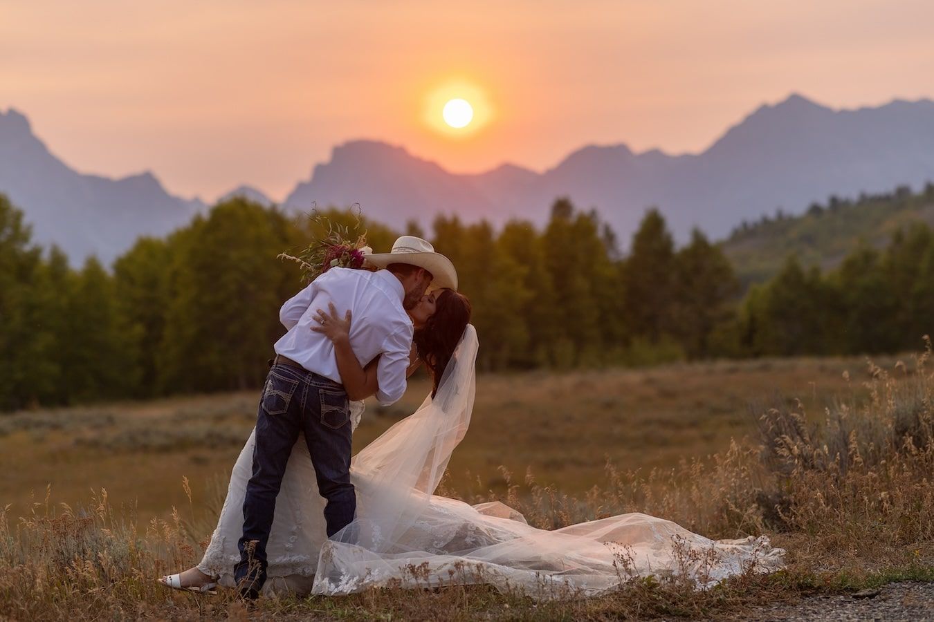 A couple embraces in a field during sunset with mountains silhouetted in the background.