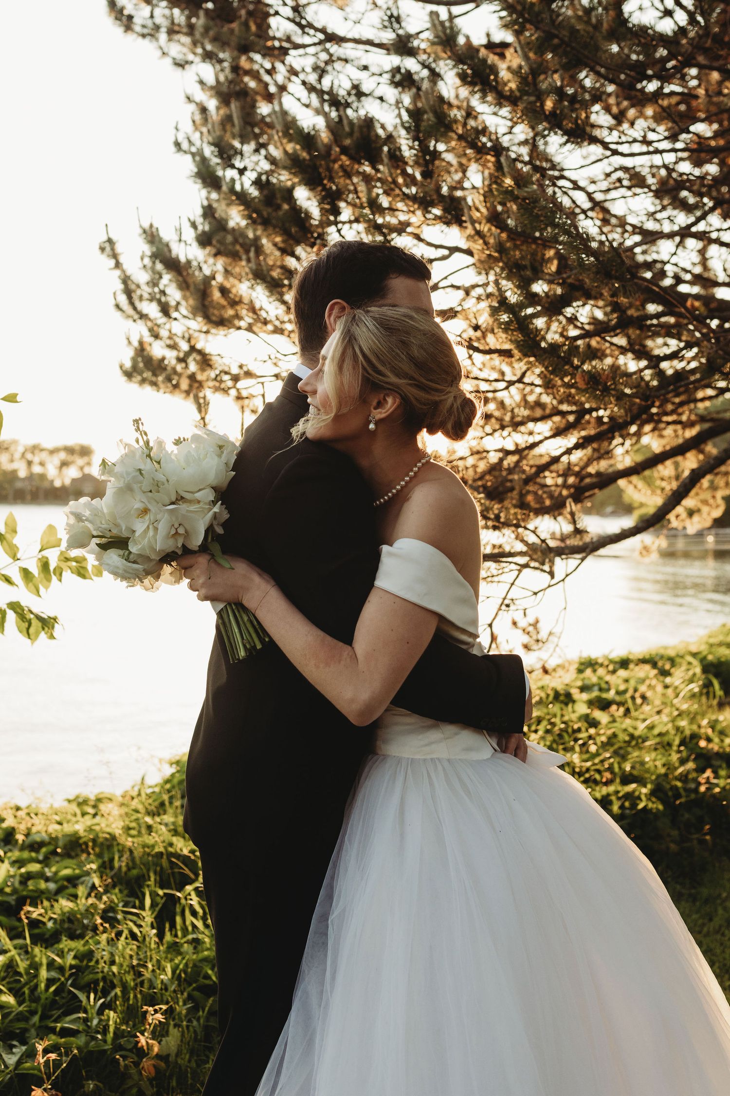 A romantic wedding couple embraces at sunset by a lake with pine trees and a white bridal bouquet in golden light.