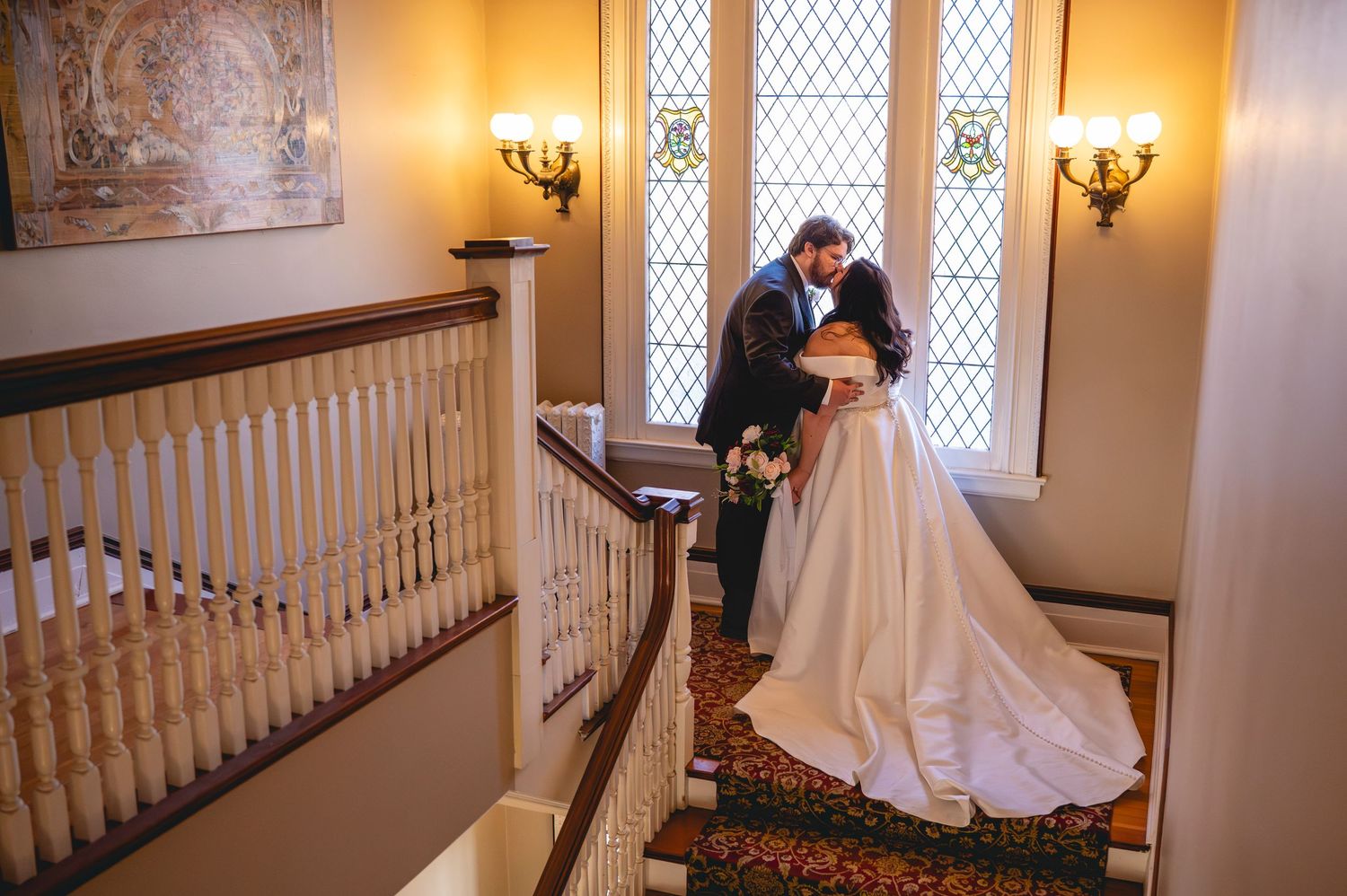 Romantic wedding couple embraces on elegant staircase in warm-lit venue with vintage windows and decor.