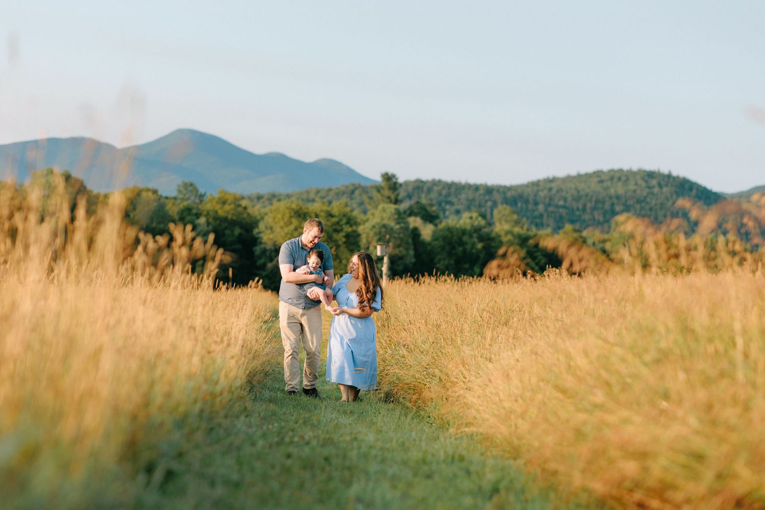 A couple stands together in a tall golden grass field with blue mountains in the distance during sunset.