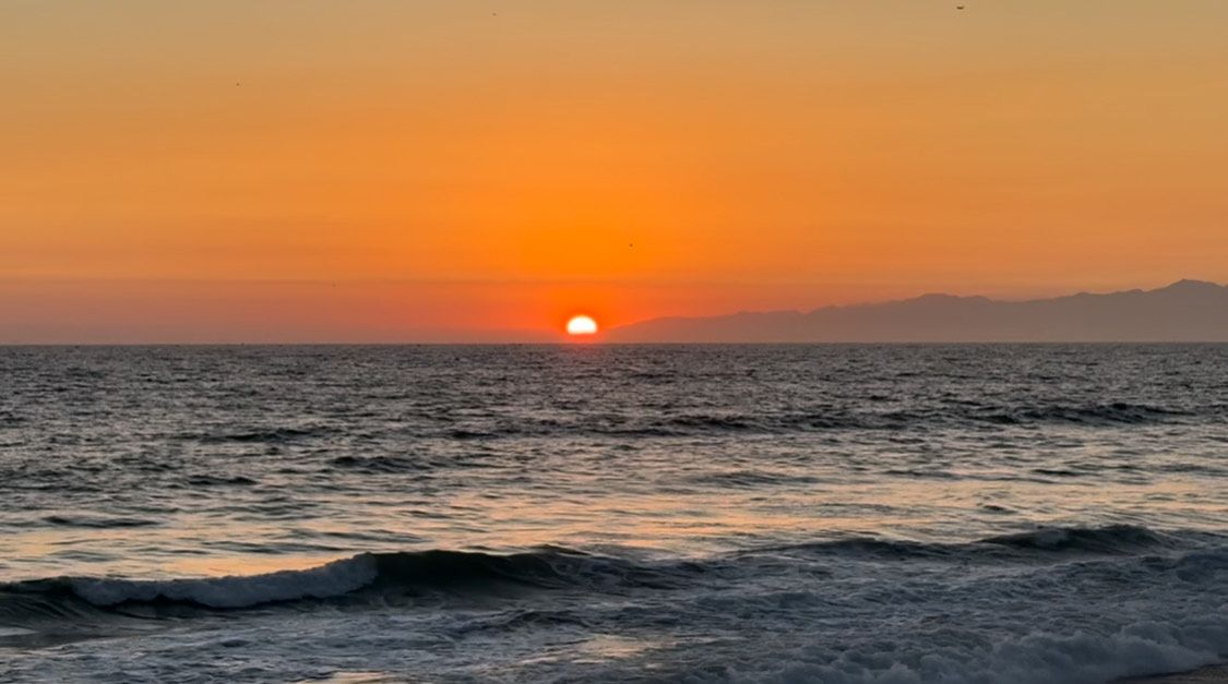 A peaceful beach landscape featuring overlapping waves crashing on shore during a vibrant sunset with mountains in distance.