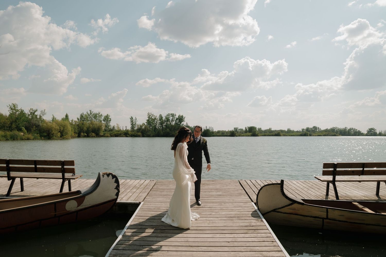 Wooden dock extends over calm lake waters surrounded by trees and blue sky on a peaceful summer day.