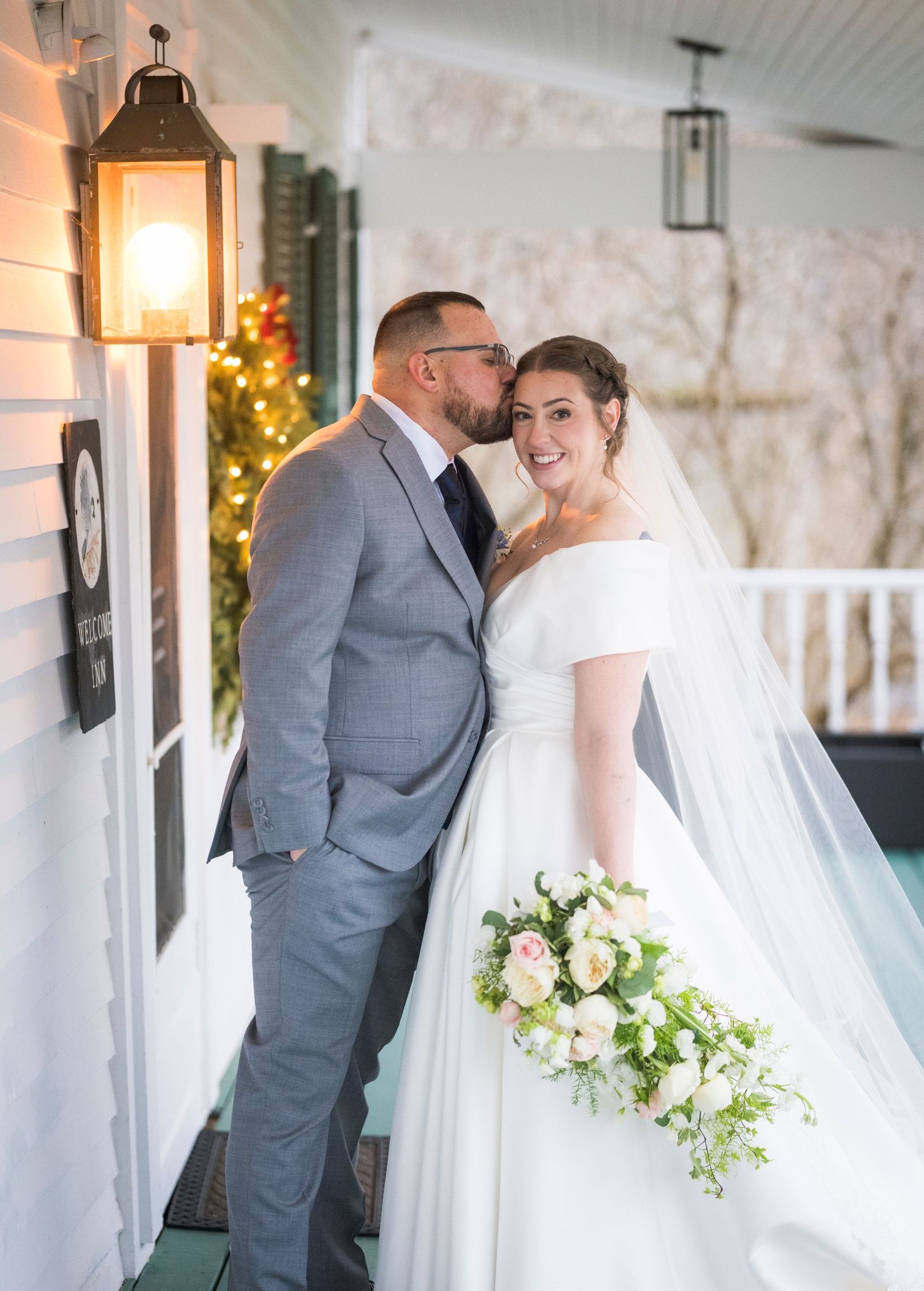 A romantic wedding moment on a porch with a glowing lantern, featuring a couple in grey suit and white gown with flowers.