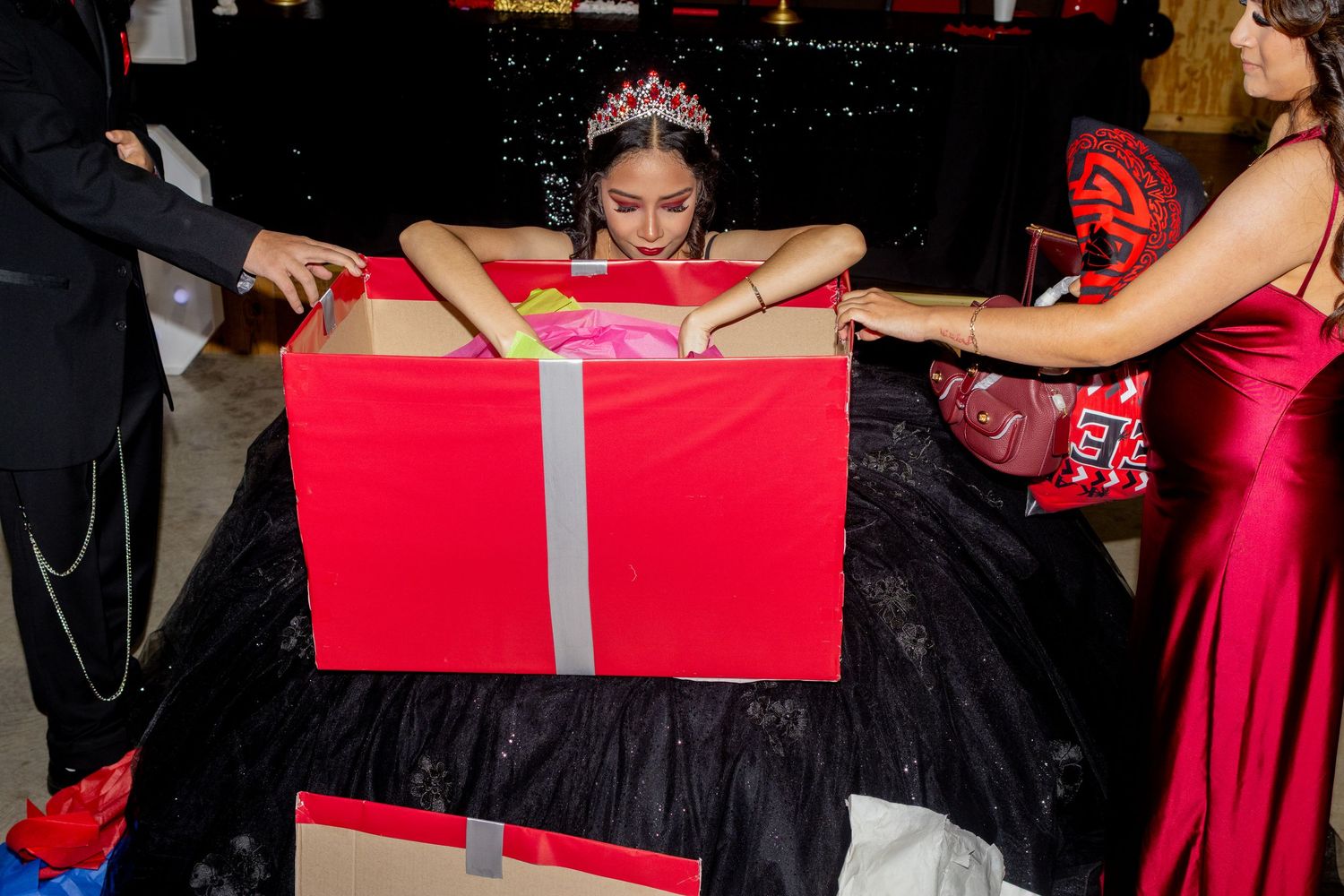 A person in a black gown poses with a large red gift box with silver ribbon at a formal event.