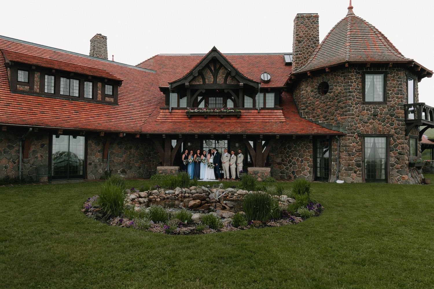 A panoramic view of a rustic stone lodge with red roof tiles and landscaped gardens set against a green lawn.