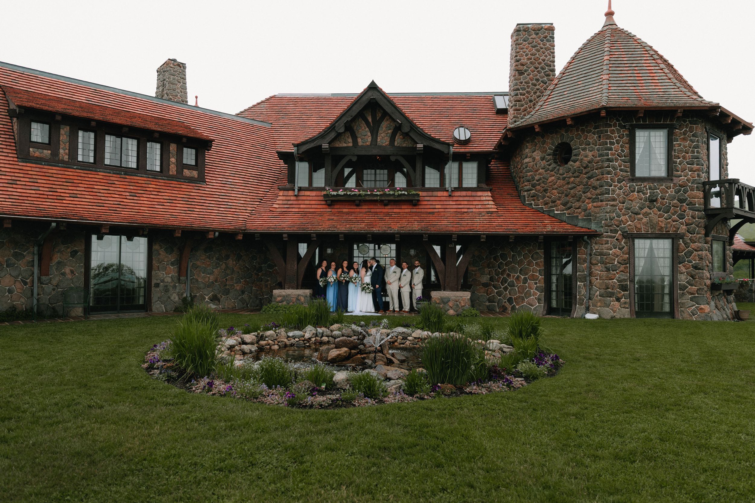 A panoramic view of a rustic stone lodge with red roof tiles and landscaped gardens set against a green lawn.