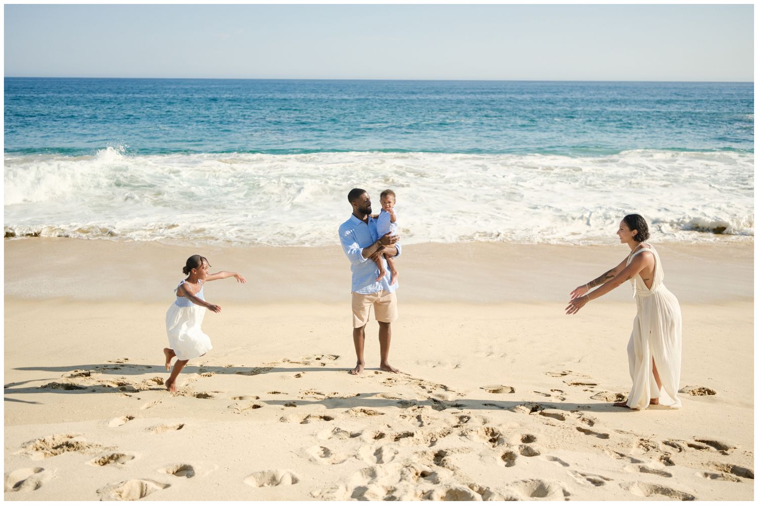 Beachgoers in white and pastel clothing gather near large rocky formations on a sandy beach during golden hour.