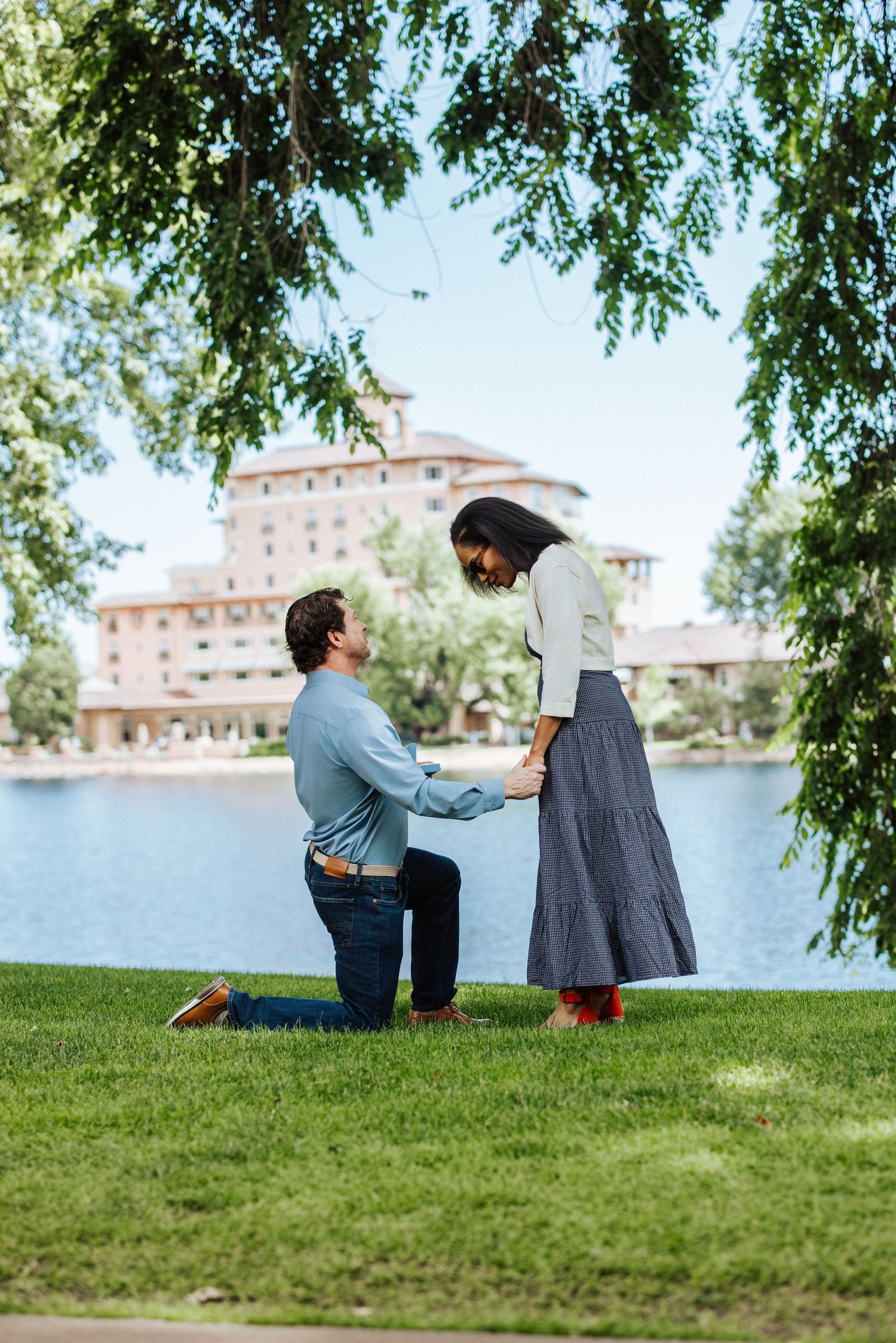 A romantic proposal scene by a lake with a historic hotel building in the background under hanging tree branches.