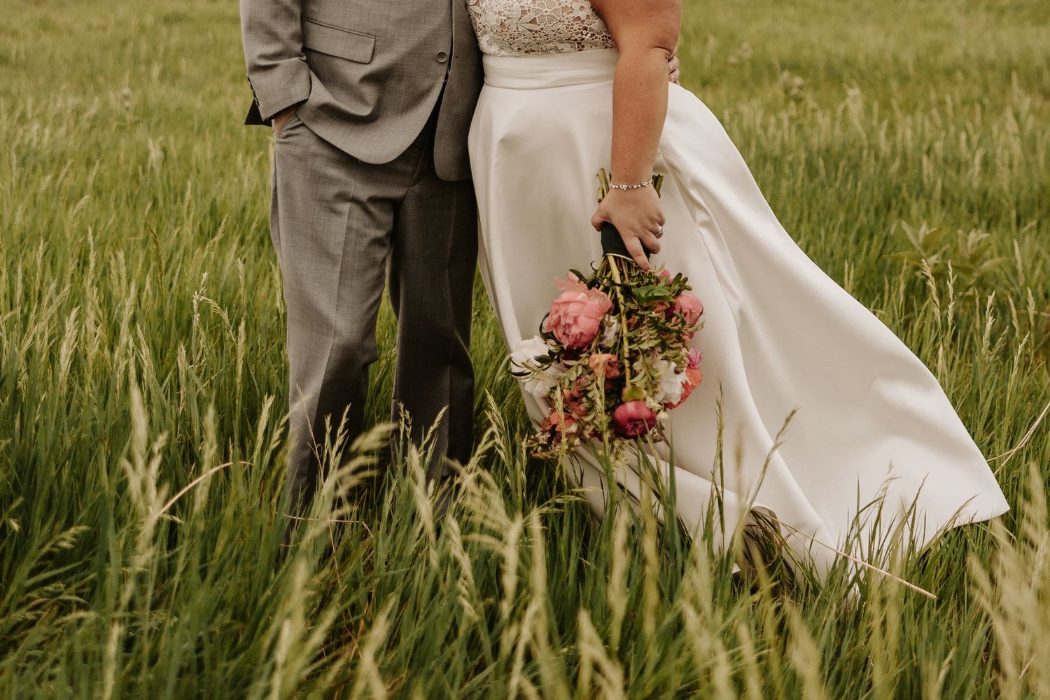 A wedding couple stands in tall grass with the bride holding a pink floral bouquet against her white dress.