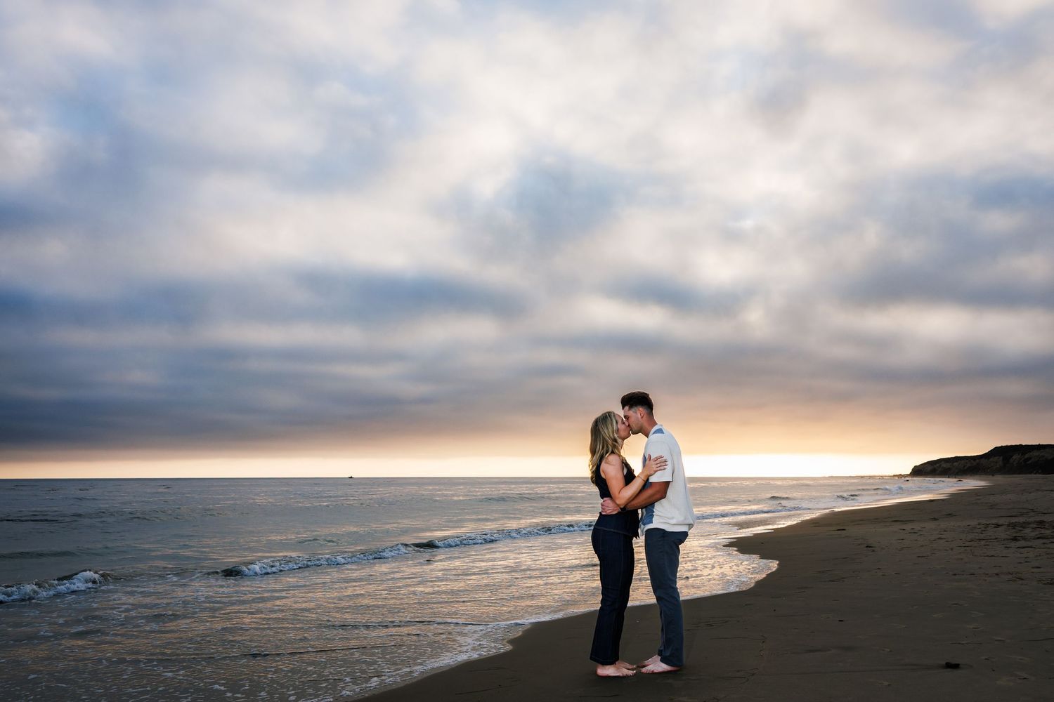 Couple embracing on a misty beach during sunset with dramatic cloudy sky.