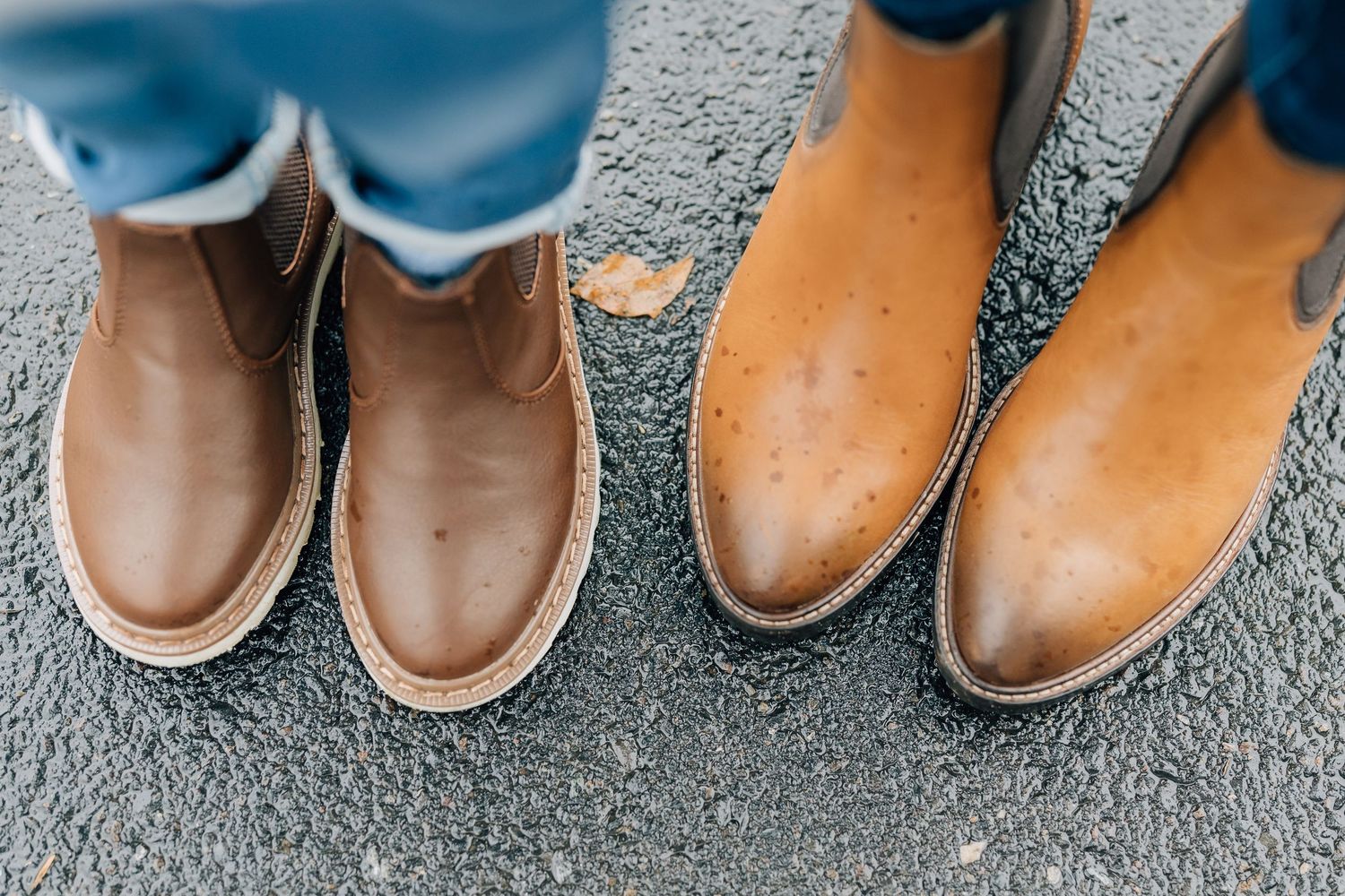 Close-up of two pairs of tan leather Chelsea boots on asphalt pavement.