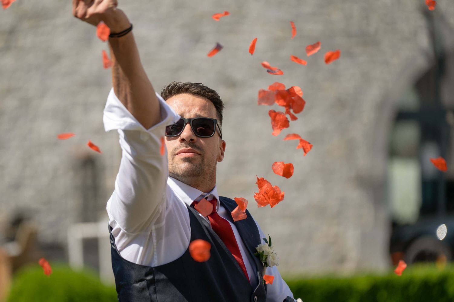 Business person in sunglasses and suit vest throws red flower petals in the air on a sunny day outside.