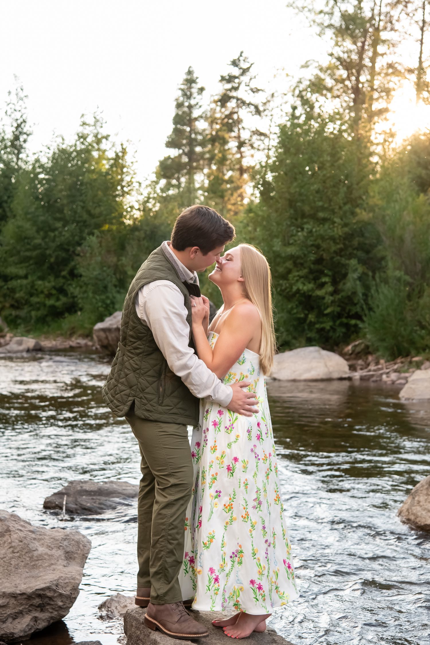 Romantic couple portrait by rushing river at sunset with pine trees in background.