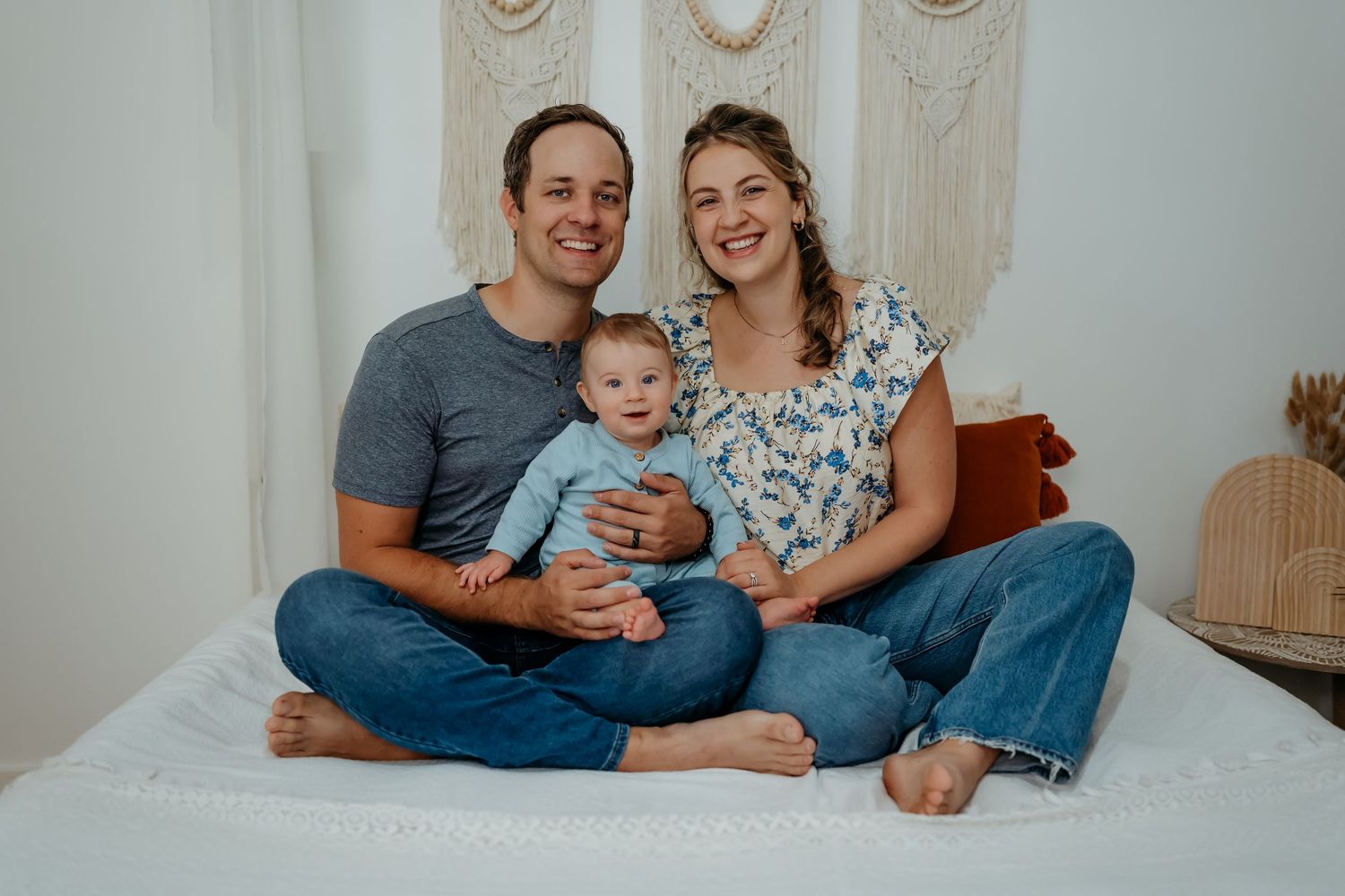 A family sits together on a white bed while sharing tender moments during an indoor photo session.