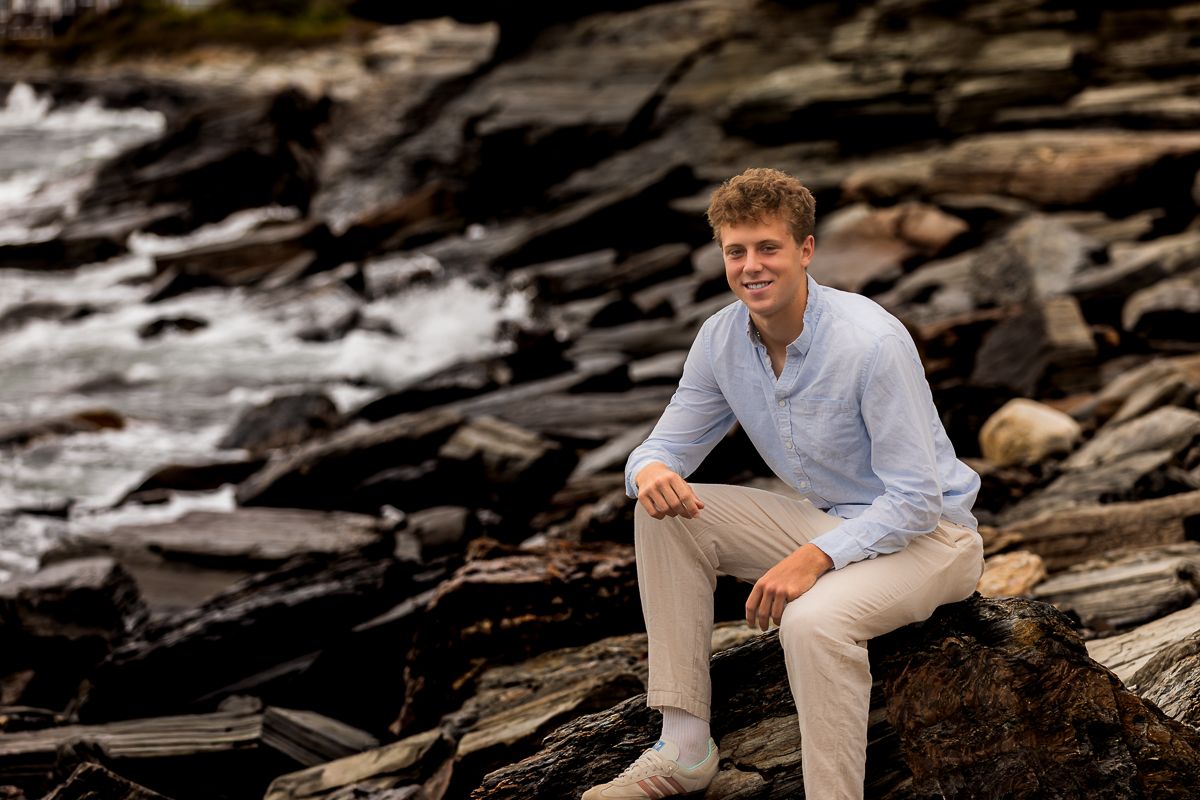 Person in blue shirt and khaki pants sitting casually on rocky coastline at sunset.