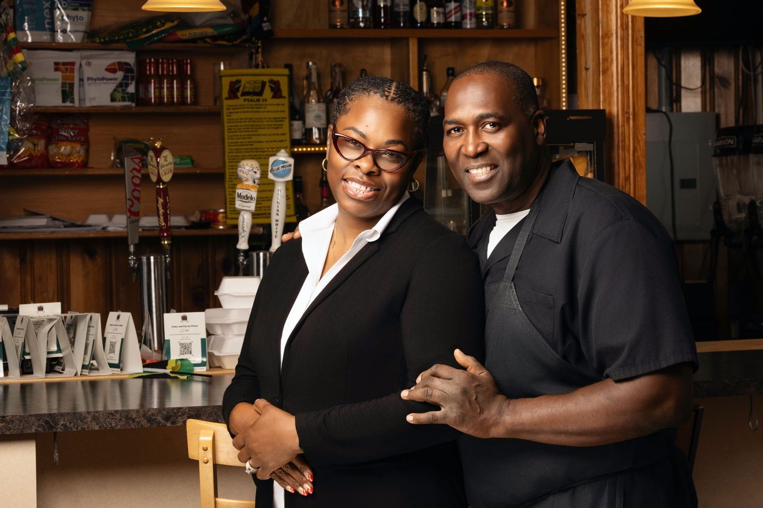 Two restaurant workers smiling together behind a wooden bar with liquor bottles visible in the background.