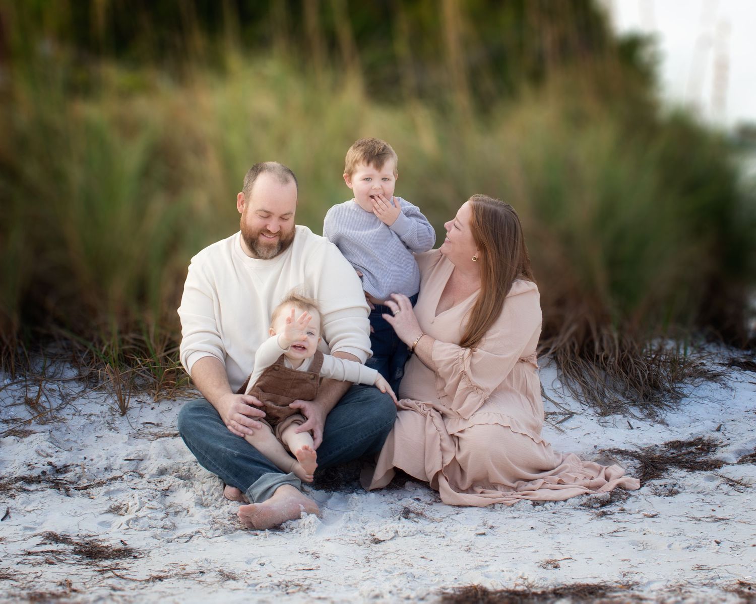 A family in neutral colored clothing sits together on a white sandy beach during a casual outdoor photo session.