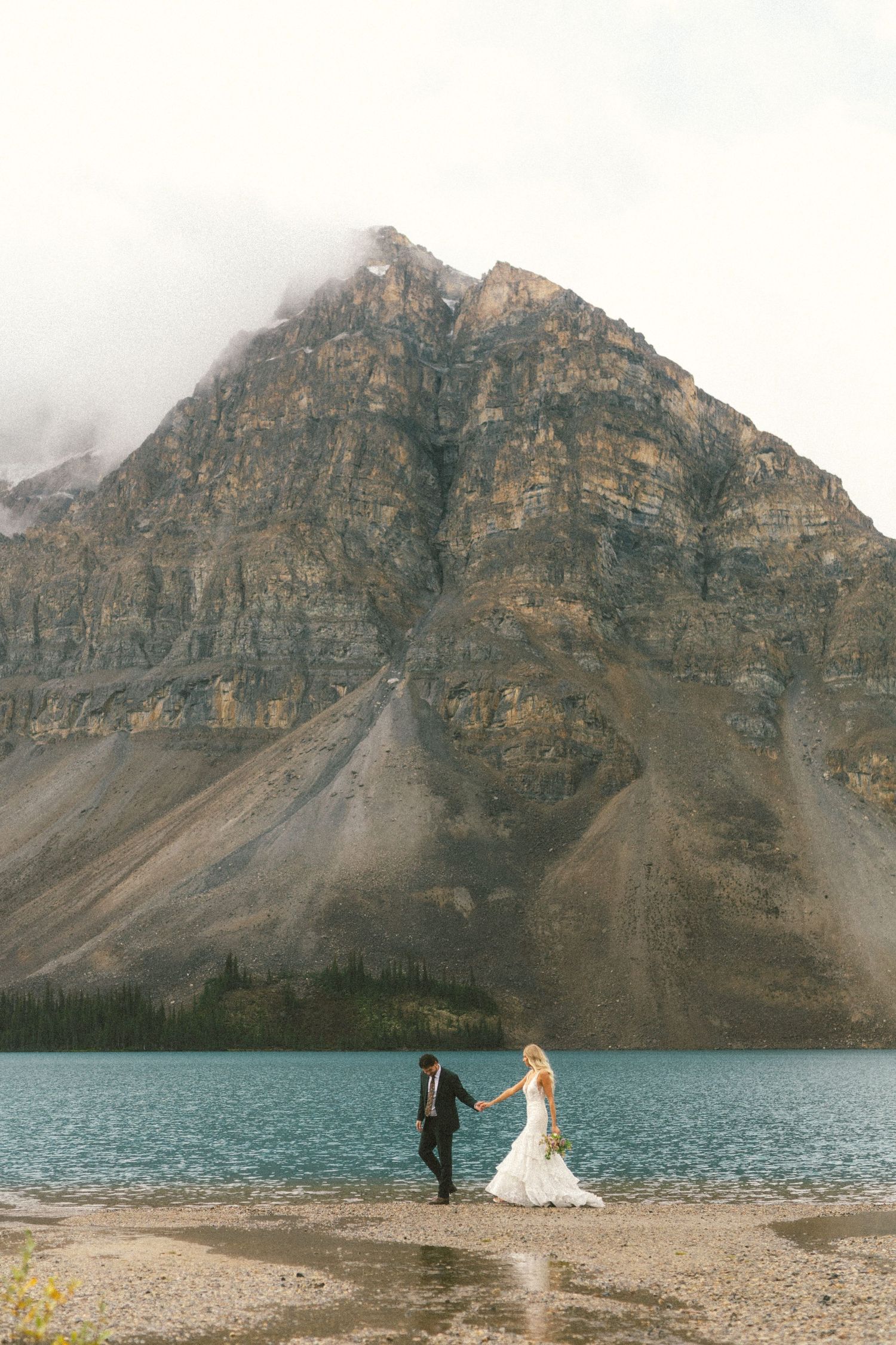 bride and groom walk along the shore of Bow Lake in Banff National Park holding hands with mountains towering above them